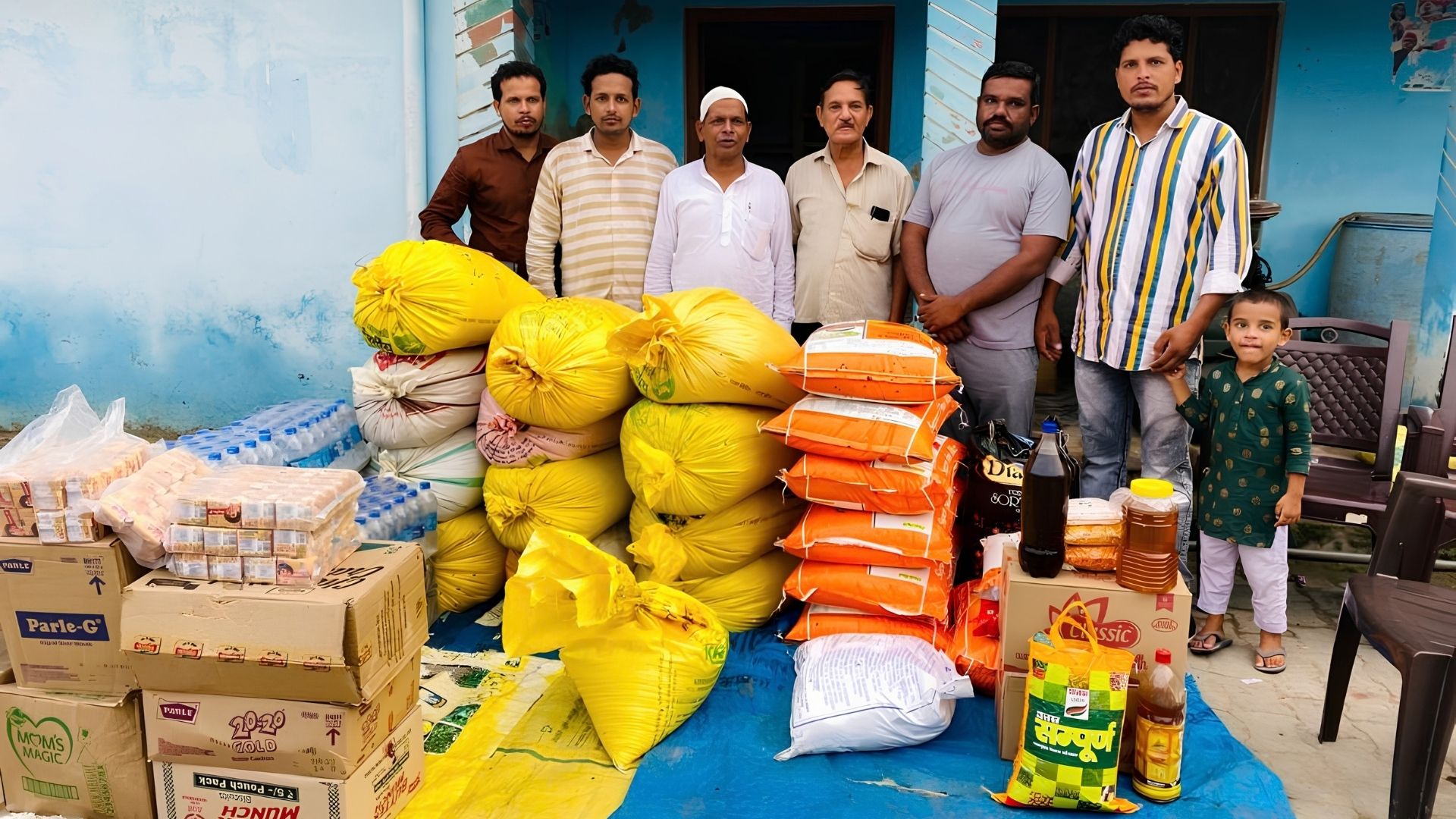 Volunteers standing behind sacks of flour, rice and pulses, cooking oil and boxes of biscuits, prepared for distribution to families.