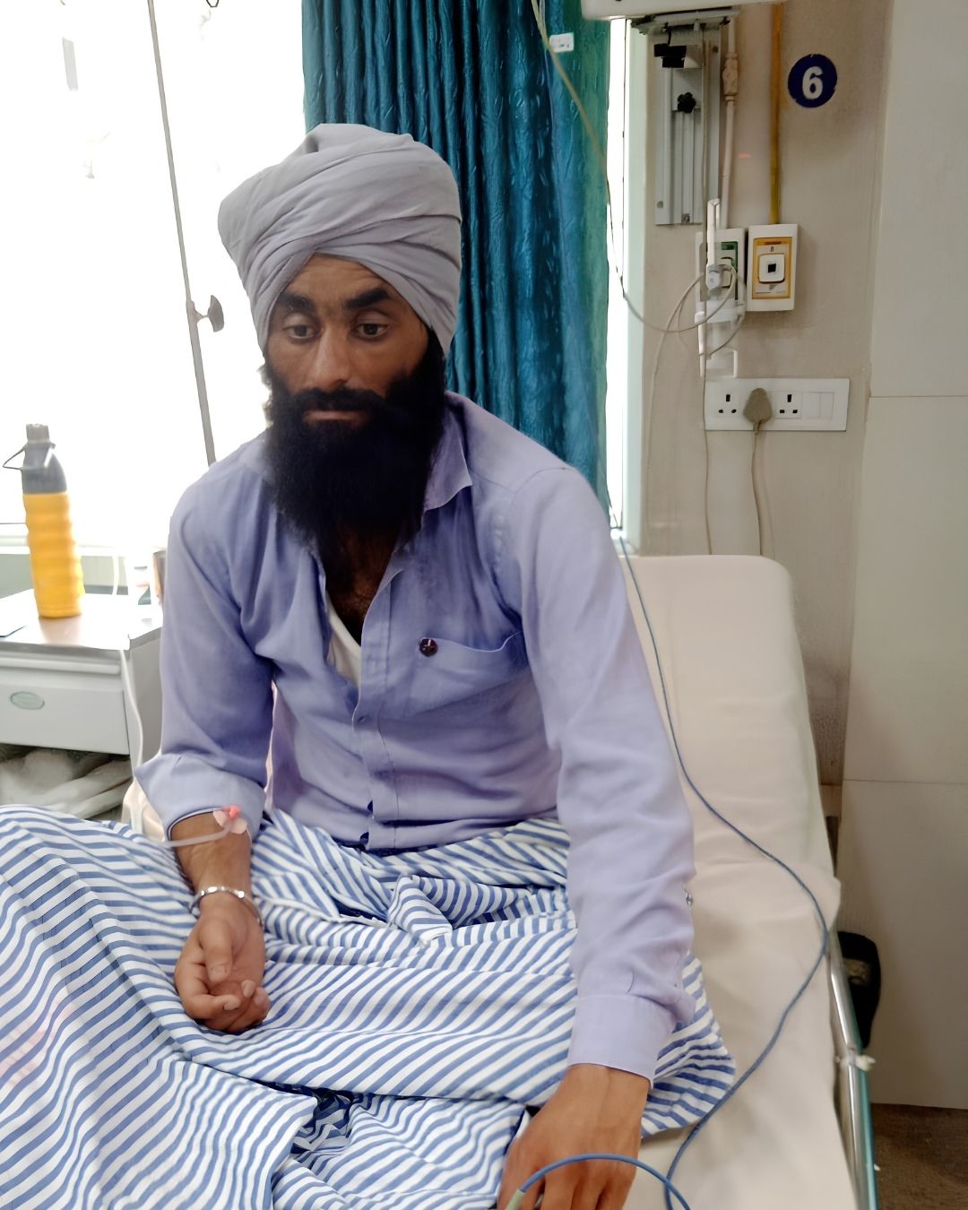 A man in a turban sitting on a hospital bed with a cannula taped to his hand, in a ward bay numbered six.