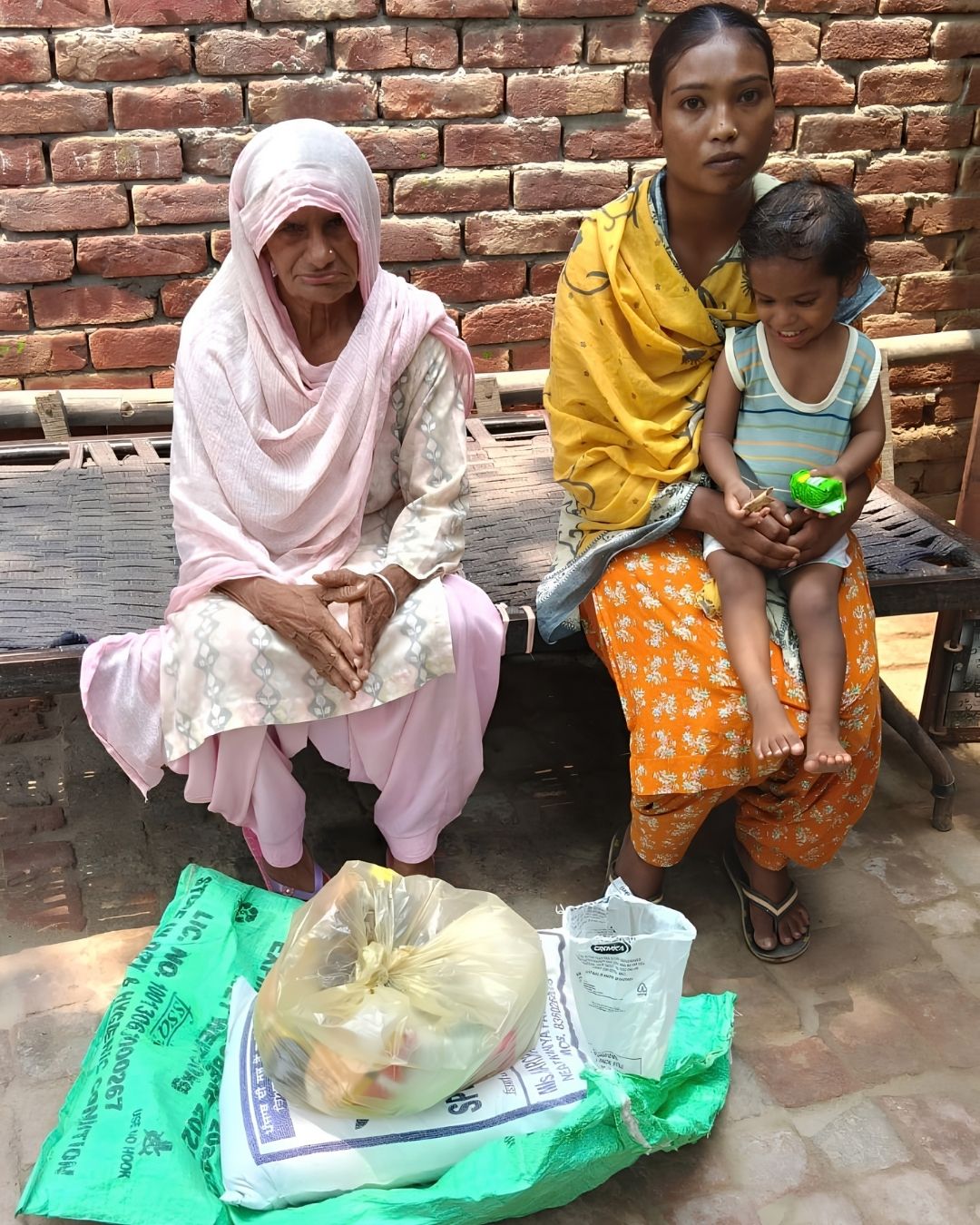 An elderly woman and a younger woman holding a small child, seated on a charpai outside a brick house, with a sack of flour and bags of provisions on the ground in front of them.