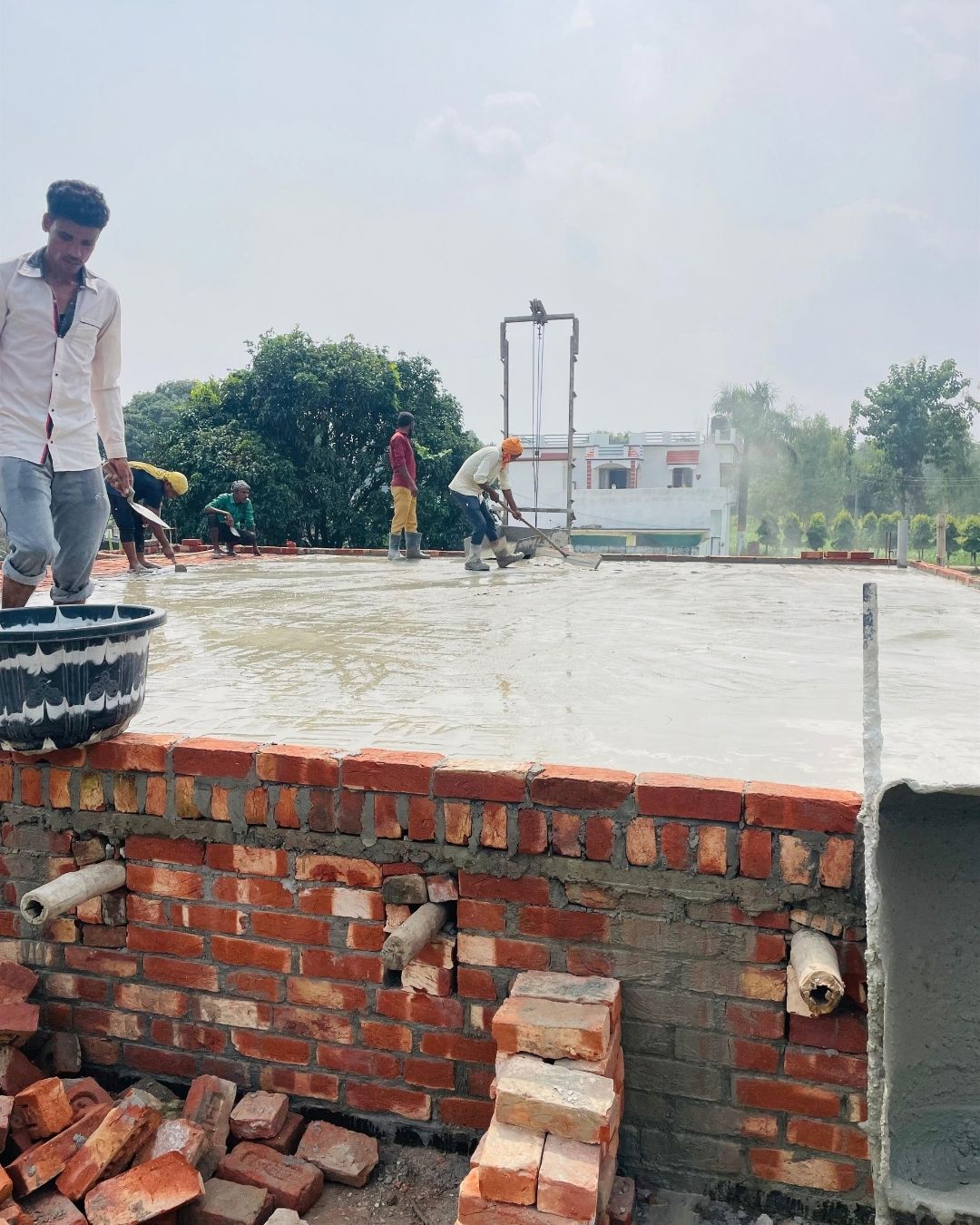 Builders spreading and levelling a freshly poured concrete roof slab on a half-built house, with stacked bricks along the parapet below.