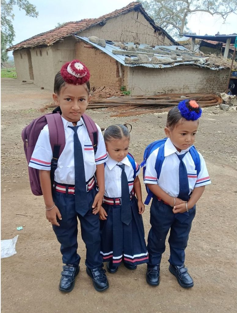 Three young children in school uniform, two carrying backpacks, standing on the bare ground outside a village home.
